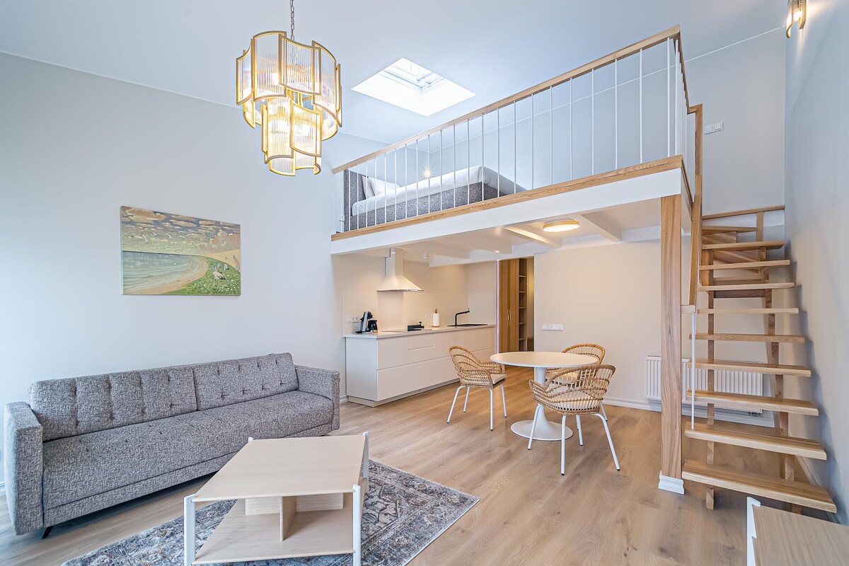 A modern loft apartment features a cozy lounge area with a gray sofa and a light-colored coffee table. A wooden staircase leads to an upper sleeping loft. The dining area includes a round table surrounded by wicker chairs. Soft natural light is provided by the skylight above.