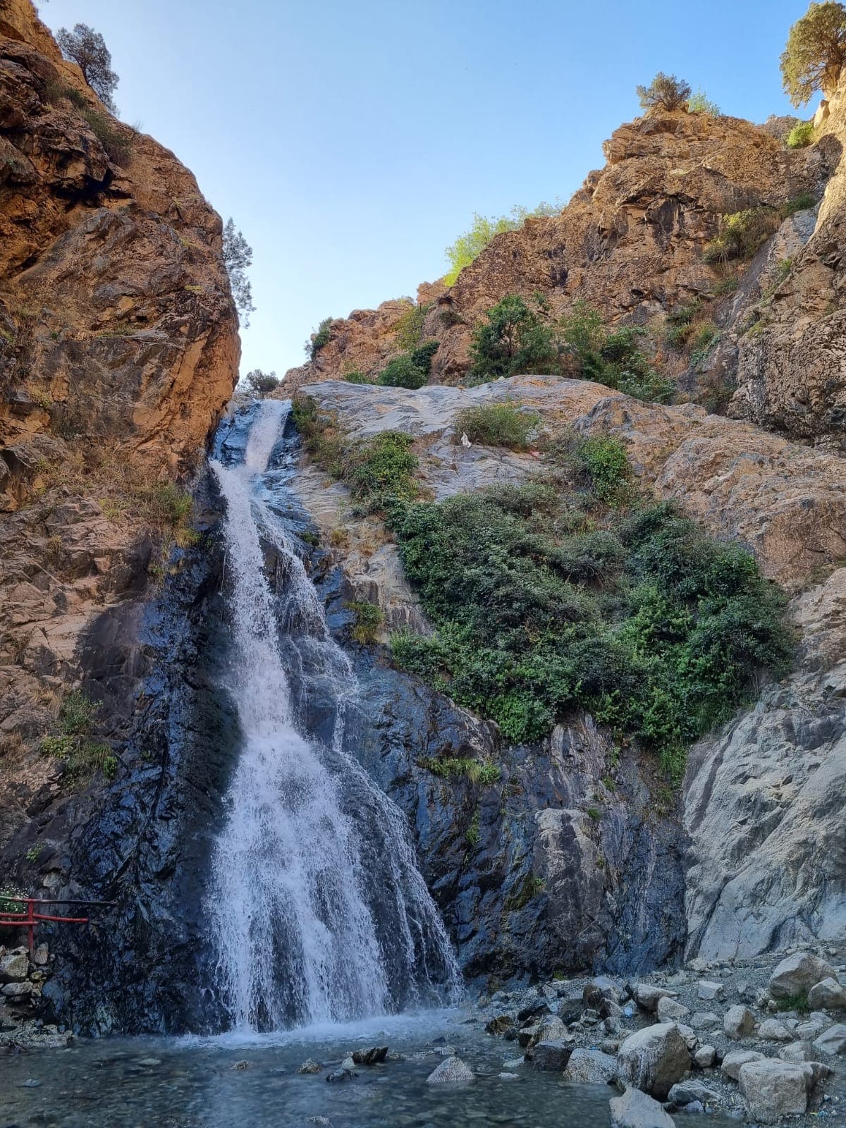A cascading waterfall descends dramatically between two rocky cliffs, surrounded by lush greenery. Sunlight illuminates the scene, highlighting the natural textures of the rocks and foliage. The base of the waterfall is visible, with water pooling over smooth stones below.