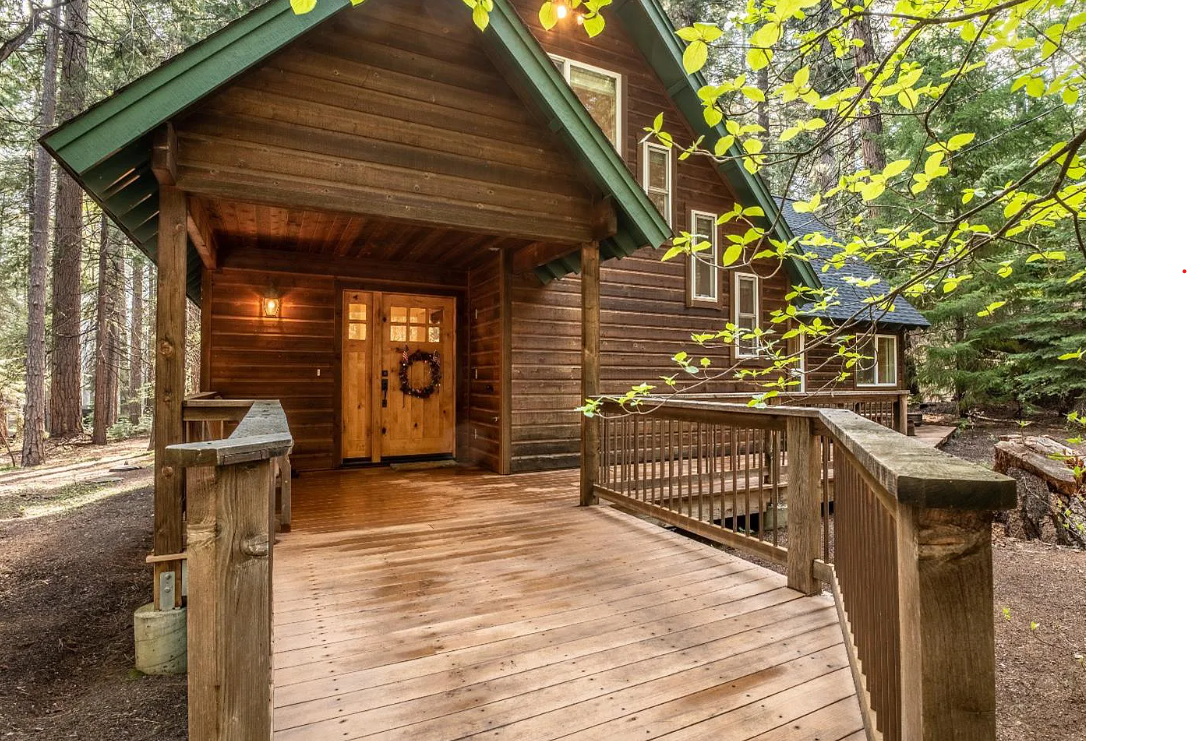 The cabin's entryway features a welcoming wooden deck, leading to double doors framed by a seasonal wreath. Surrounding trees create a peaceful wooded atmosphere, while green accents on the roof complement the natural setting.