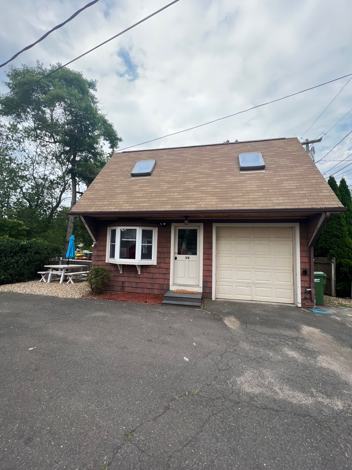 The exterior of a newly renovated cottage features a sloped roof with skylights. A front porch welcomes visitors, and a driveway leads to a garage. Outdoor seating is provided with a picnic table and an umbrella visible in the background amid a landscaped area.
