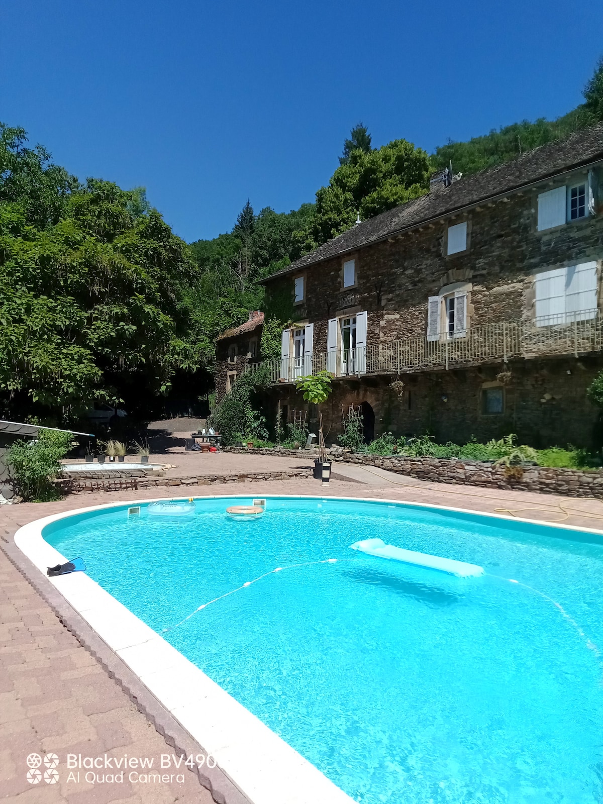 An inviting pool is centered in the image, featuring clear blue water. The surrounding area includes a stone structure with multiple windows and a lush green backdrop of trees. A patio area is visible with outdoor seating, creating a peaceful outdoor environment.