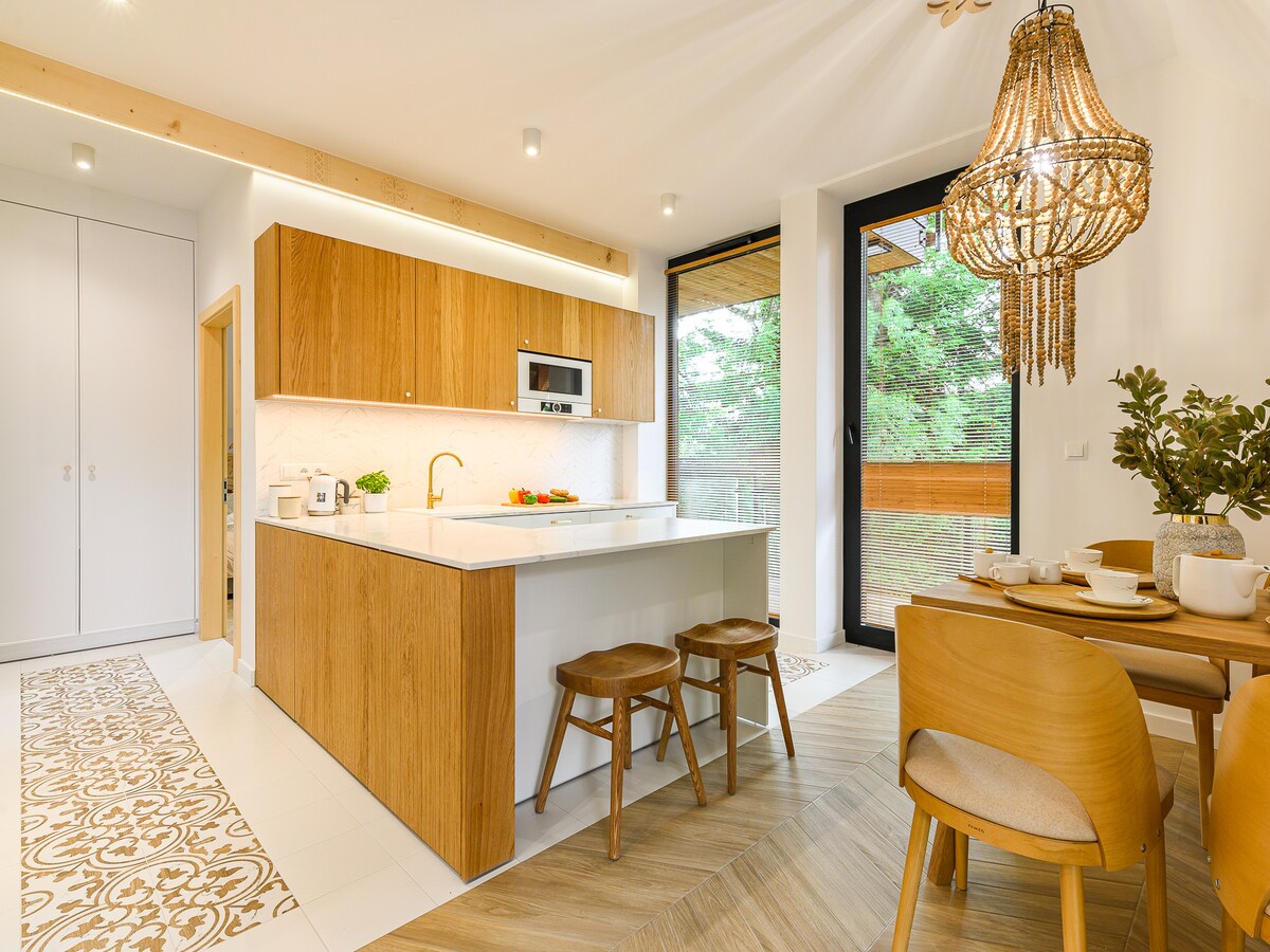 A modern kitchen features wooden cabinetry and a central island with seating for three. Natural light fills the space through large glass doors that open to a view of greenery. Decorative elements include a statement chandelier and a dining area with elegant table settings.