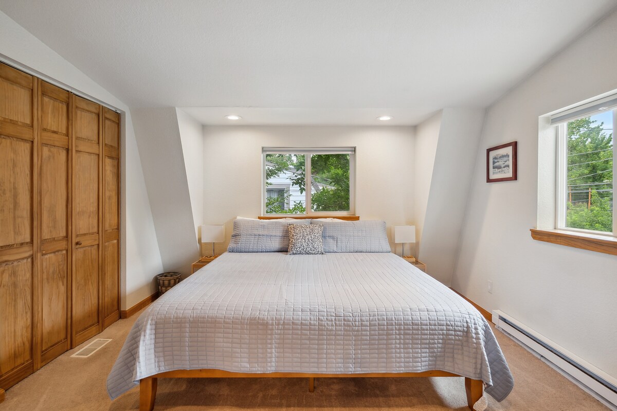 The bedroom features a spacious bed covered with a light quilt and a decorative pillow. Natural light enters through a window flanked by two bedside lamps. A large closet with wooden doors is visible, complementing the neutral wall tones and soft carpeting.