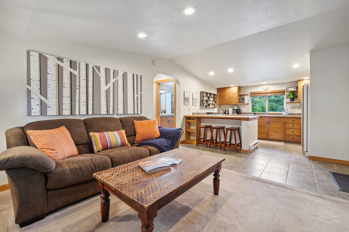 A spacious living area showcases a comfortable brown sofa with accent pillows, paired with a wooden coffee table. The kitchen area features wooden cabinets and a breakfast bar with stools, while large windows provide ample natural light.