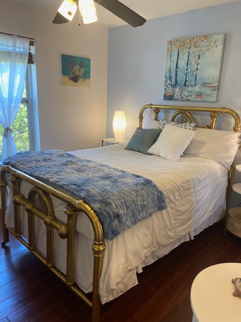 A cozy bedroom features an antique brass bed with soft linens and decorative pillows. A light blue throw is draped over the foot of the bed. Natural light enters through a window adorned with sheer curtains, and art hangs above the bed, creating a calm atmosphere.