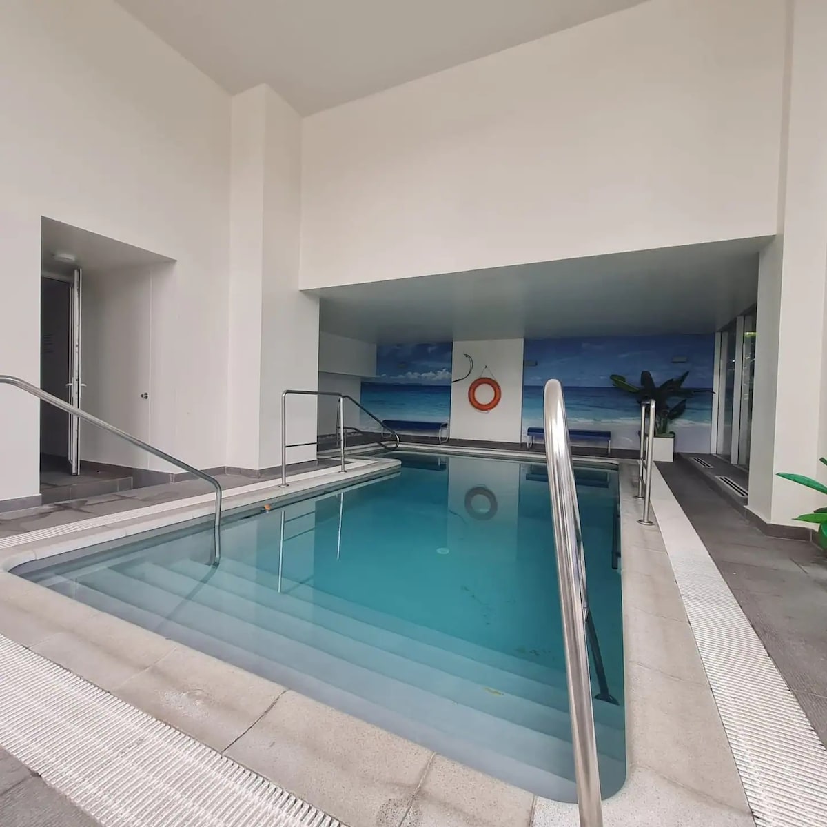 An indoor heated pool is featured with smooth tiles surrounding it. Stainless steel railings provide access, and a mural depicting a beach scene is displayed on the wall. Natural light enters through large windows, enhancing the tranquil environment.