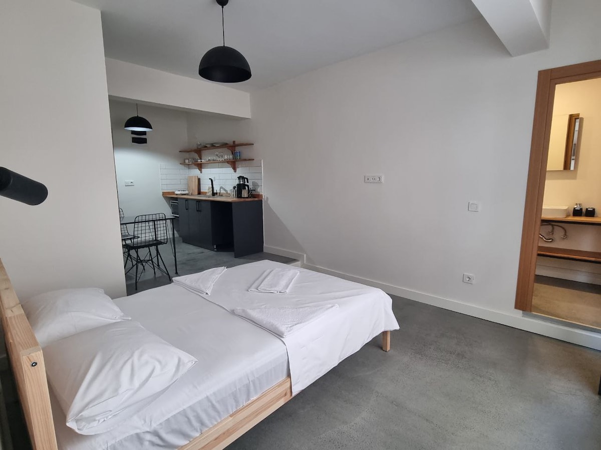 An airy bedroom features a minimalist design with a spacious bed dressed in white linens. A sleek kitchenette is visible in the background, showcasing modern appliances and open shelving. Natural light enhances the neutral tones of the space, contributing to a calm ambiance.