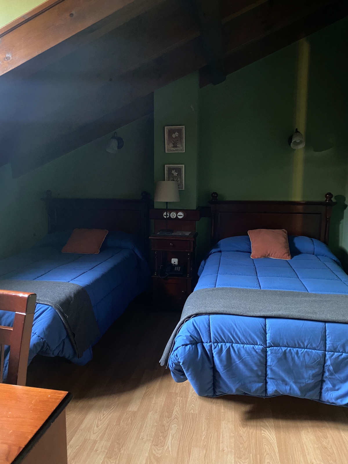 A cozy room is furnished with two beds, each covered in blue bedding. A small bedside table is placed between the beds, featuring a lamp and framed artwork on the walls. Slanted ceilings add character to the space, complemented by wooden beams.