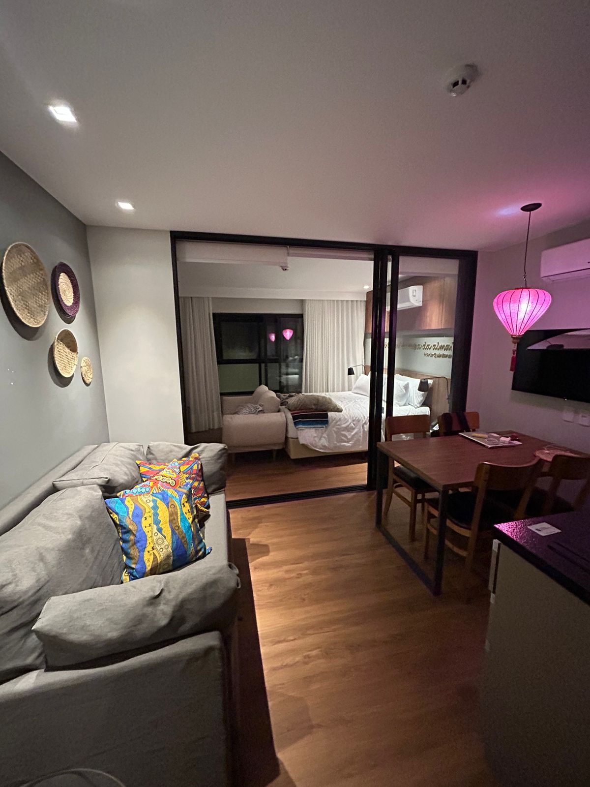 A stylish living area features a light gray sofa adorned with decorative pillows, positioned beside a wooden dining table and chairs. A soft glow from a hanging light fixture enhances the ambiance, while art pieces are displayed on the wall. Glass panels separate the space from the bedroom.