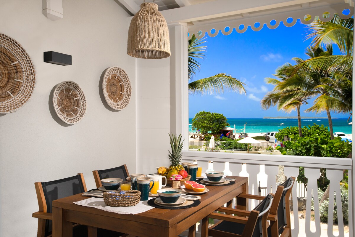 A dining area features a wooden table set for a meal, surrounded by chairs. Decorative woven plates adorn the wall. Through the open balcony, a view of a turquoise sea and palm trees creates a scenic backdrop.