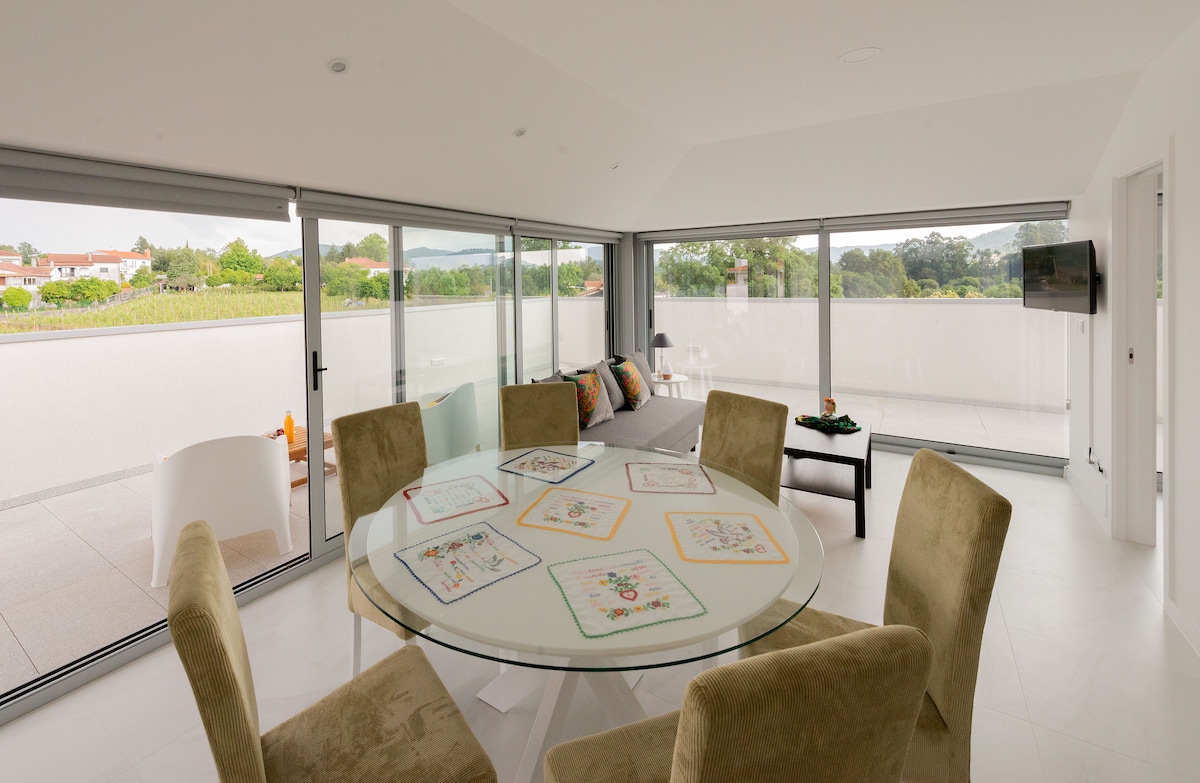 A modern dining area features a round glass table surrounded by plush, neutral-colored chairs. Large sliding glass doors offer views of the surrounding landscape and allow natural light to fill the space. A comfortable seating area with soft cushions is visible in the background.
