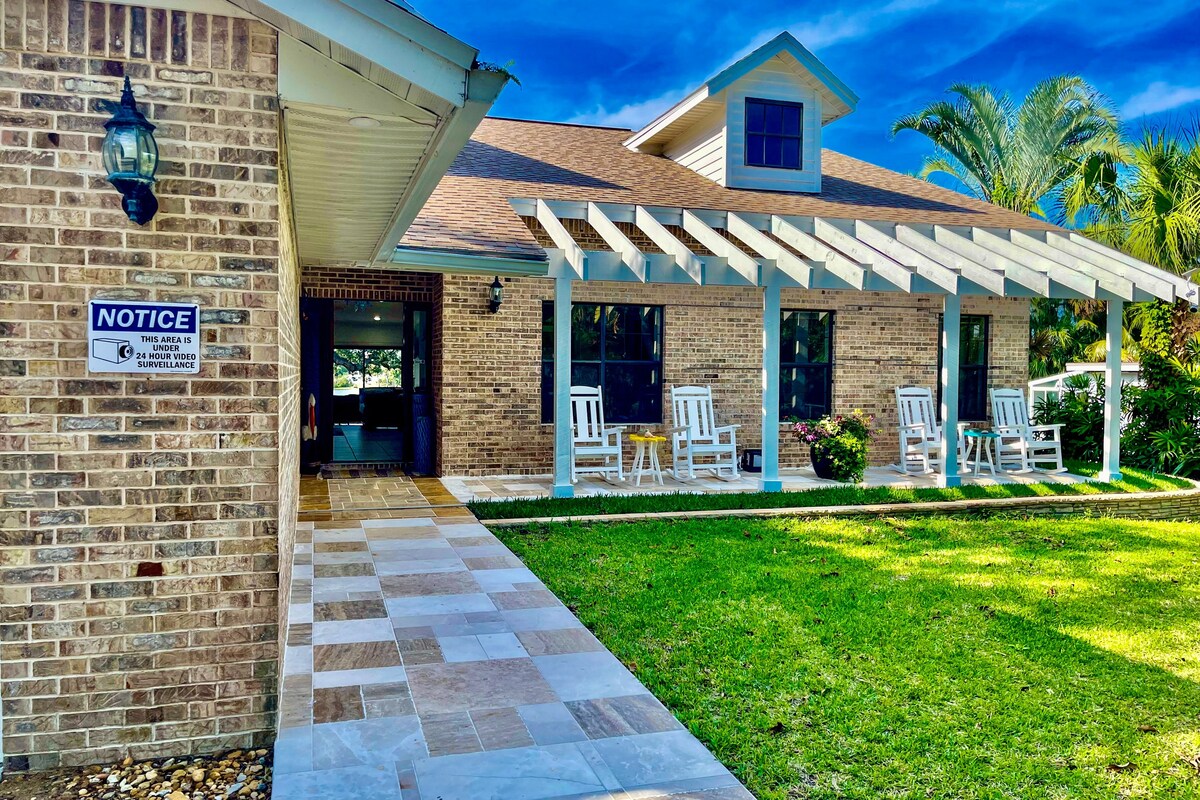 A brick house features a spacious front porch with white rocking chairs, inviting relaxation. The well-manicured lawn surrounds the entrance, while lush palm trees provide a tropical ambiance. A pathway of large stone tiles leads to the front door, complementing the inviting exterior.