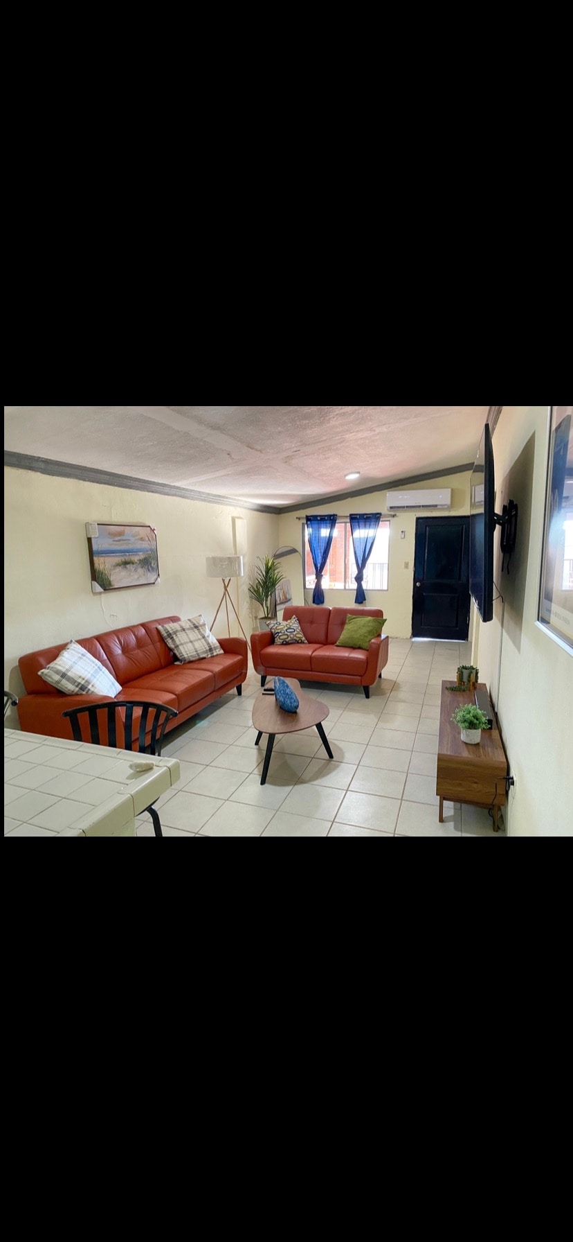 A cozy living area features two comfortable sofas, one in orange and another in green. A coffee table sits in front of the sofas, with a decorative plant nearby. Natural light enters through a sliding door, enhancing the inviting atmosphere.