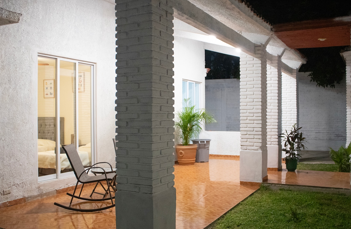 A well-lit patio area is framed by white columns, offering a view of gardens. A rocking chair is positioned on the tiled floor, and large glass doors provide access to the bedrooms inside. Lush plants are visible, enhancing the outdoor space.
