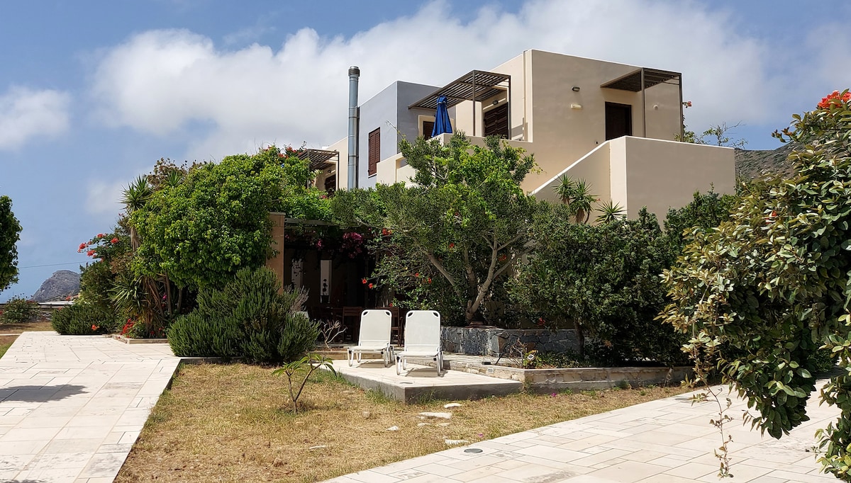 The exterior of the apartment complex displays a two-storey structure surrounded by well-maintained gardens with various flowers and shrubs. A paved pathway leads through the grass towards the entrance, where two lounge chairs are positioned for relaxation under a partly cloudy sky.