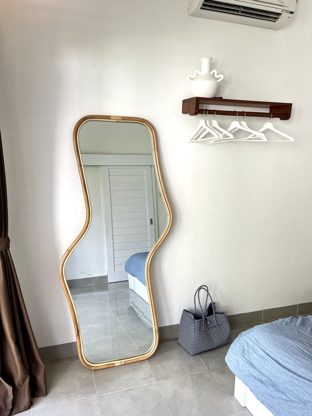 A tall, curved mirror leans against the wall, reflecting the spacious interior. Above, a wooden shelf holds several hangers, while a stylish vase adds a decorative touch. A cozy bed is partially visible, accompanied by a woven bag placed on the floor.
