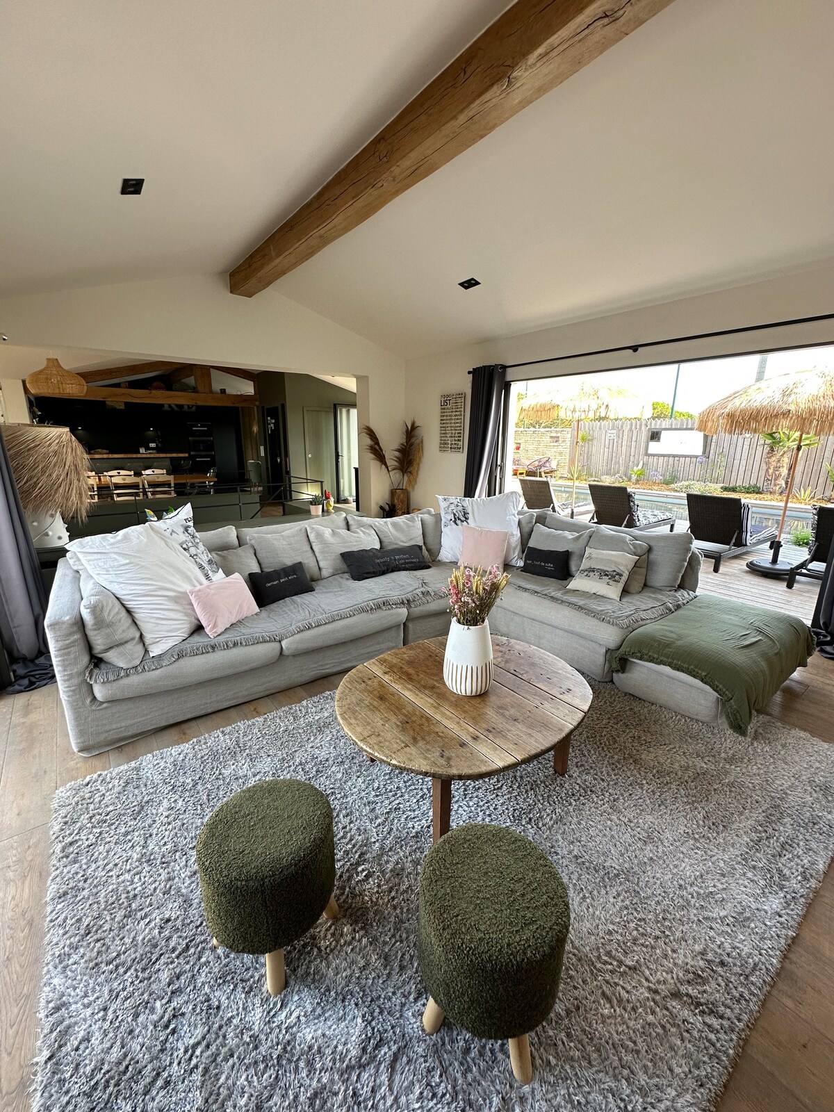 A spacious living area is highlighted by a large sectional sofa adorned with pillows. A round wooden table sits at the center, surrounded by soft rugs. Glass doors open to an outdoor dining space, creating a seamless transition to the patio.