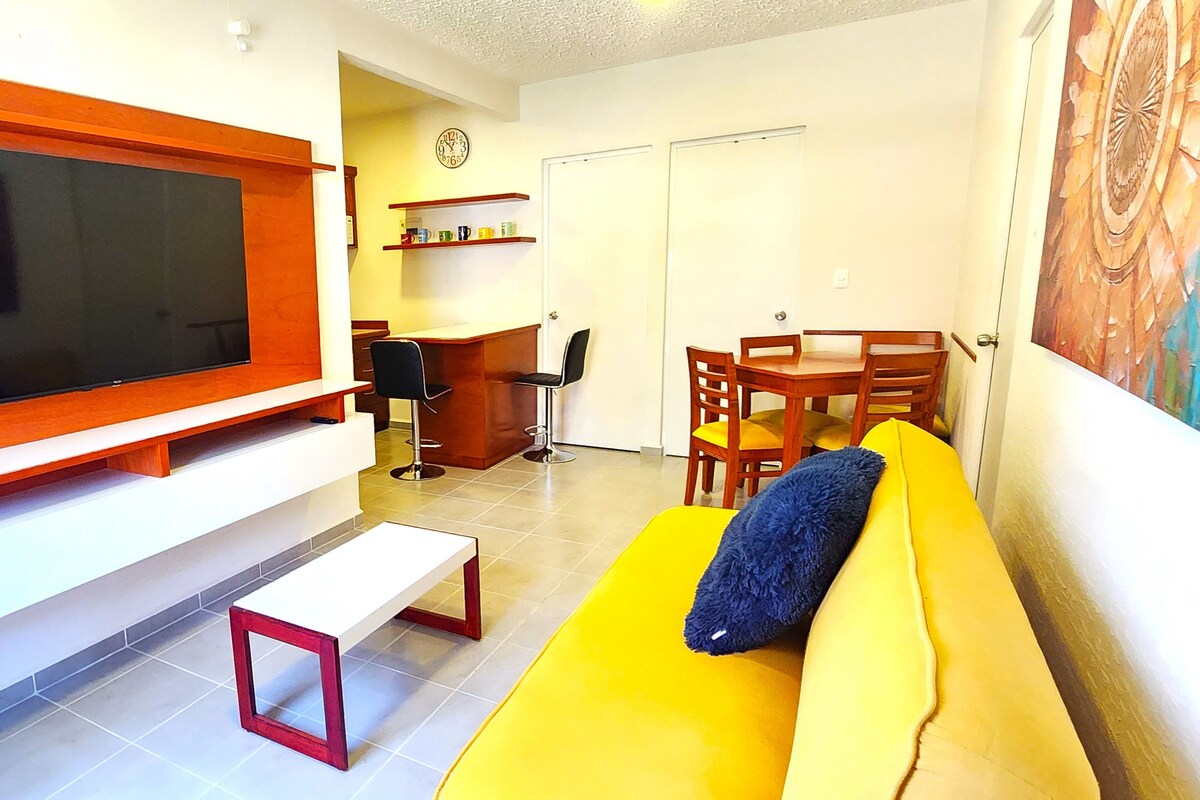 A living area features a vibrant yellow sofa paired with a white coffee table. A wall-mounted TV is positioned above a sleek wooden shelf. In the background, a small dining table with four wooden chairs and a work desk with black chairs are visible, enhancing the space's functionality.