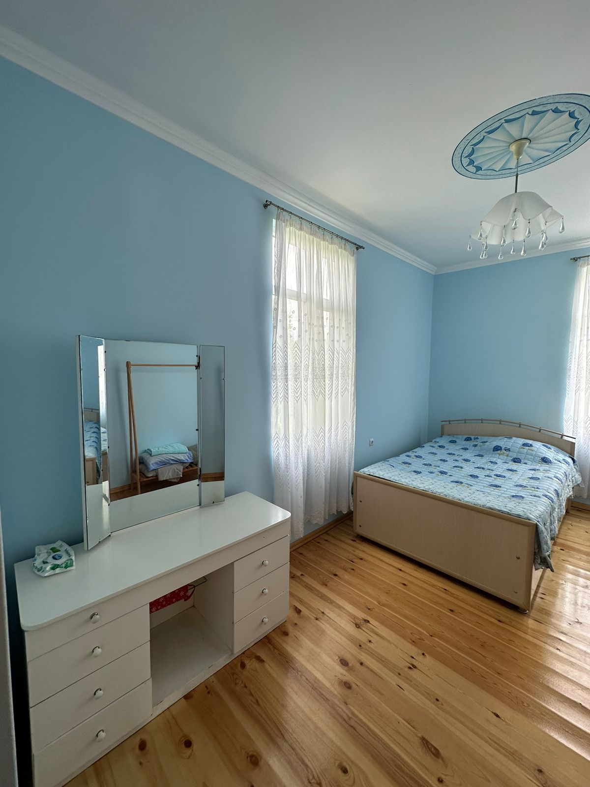 A light blue bedroom features a spacious bed with a patterned duvet and a matching pillow. A white vanity with multiple drawers is placed against one wall, and natural light filters through sheer curtains hung by two windows.