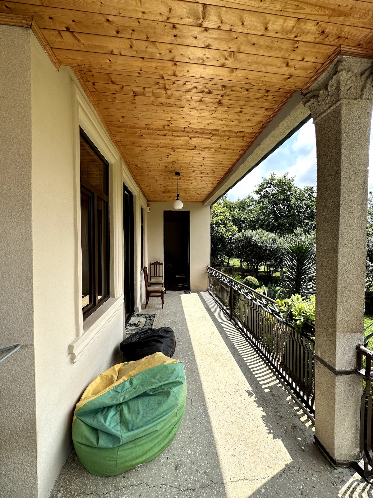 A covered porch is visible, featuring a wooden ceiling and decorative columns. Comfortable bean bags are placed alongside a wooden chair. Sunlight creates a warm ambiance, while greenery can be seen in the distance, contributing to a serene outdoor setting.