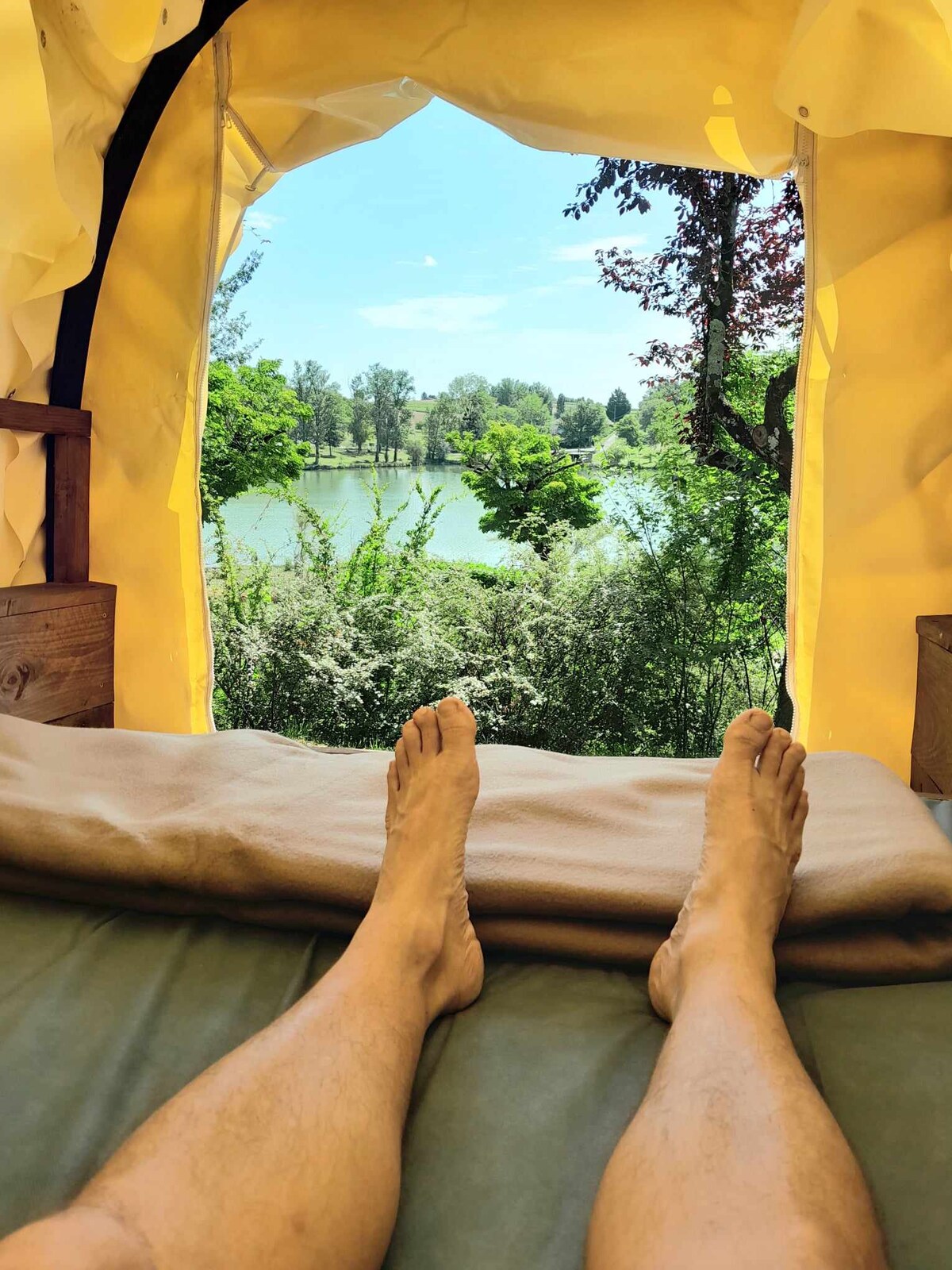 A view from an indoor space features an open window framing a scenic lake surrounded by lush greenery. Sunlight illuminates the area, enhancing the calming natural atmosphere. In the foreground, bare feet rest on a simple bed with soft bedding.