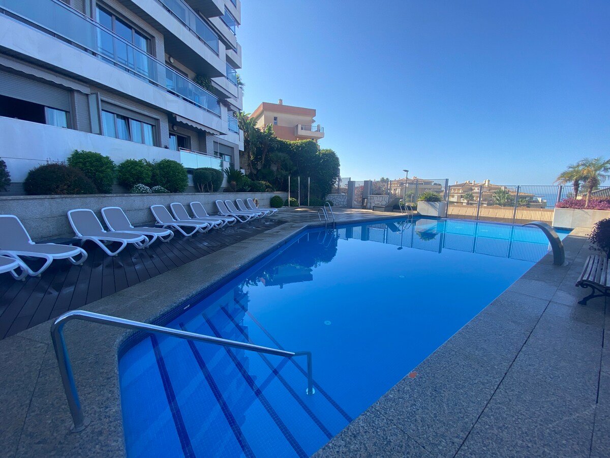 A clear blue pool is surrounded by a spacious terrace with multiple white lounge chairs. Lush greenery and palm trees provide a serene backdrop. The building's modern architecture is visible in the background, enhancing the inviting atmosphere of the outdoor space.
