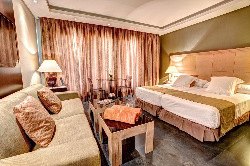 The room features two individual beds positioned beside a cozy sofa, accompanied by a coffee table. Large windows are adorned with flowing curtains, allowing natural light to fill the space. The modern decor includes neutral tones, contributing to a sophisticated atmosphere.