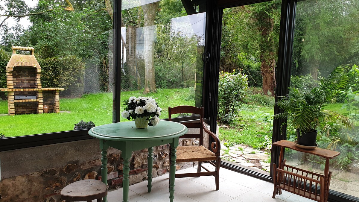 A sunlit conservatory features a round green table with a vase of flowers, complemented by a wooden chair. Large glass panels provide a view of the lush garden, showing greenery and a stone path. A small wooden cradle adds to the inviting decor.
