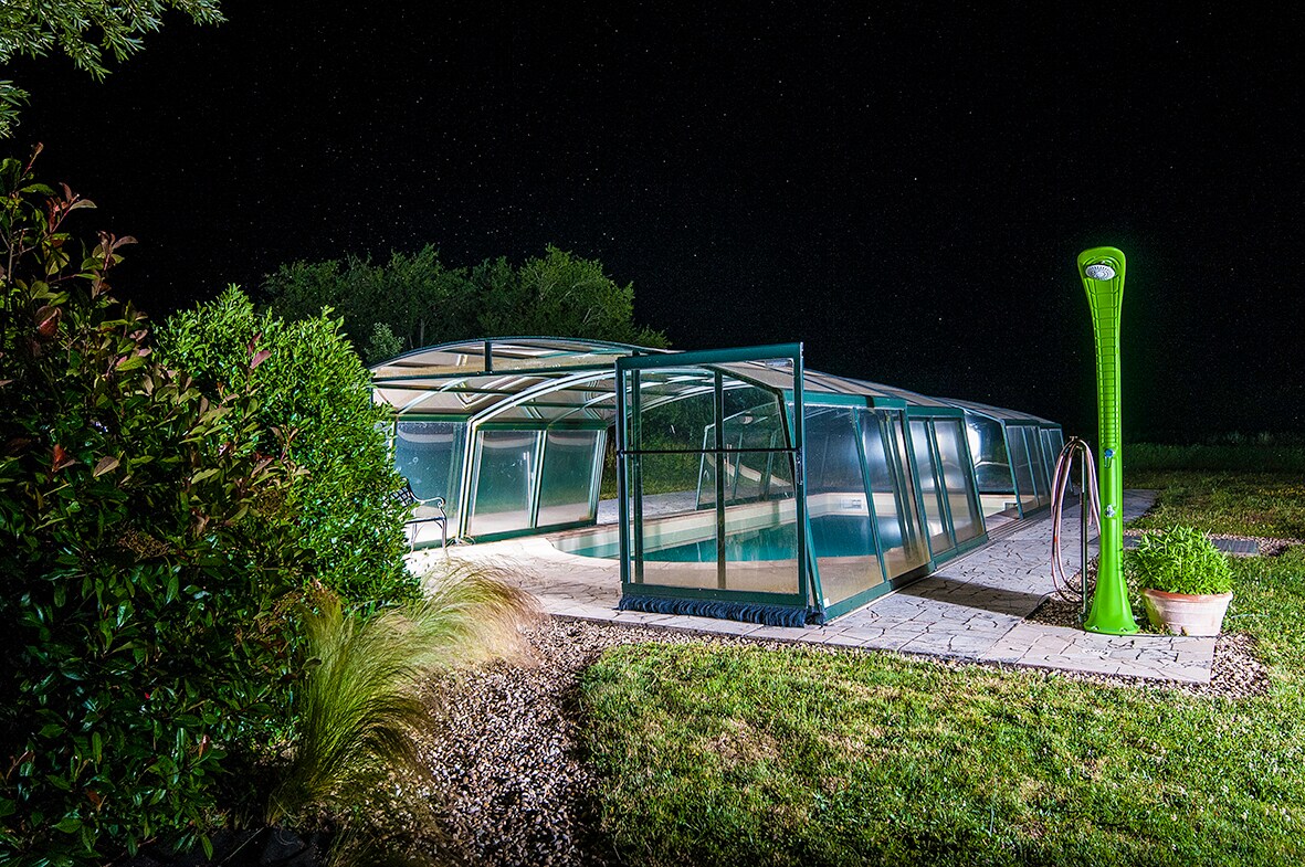 A serene outdoor space features a covered saltwater pool, illuminated under a starlit sky. Surrounding greenery adds a touch of nature, while a modern outdoor shower is positioned nearby for convenience. Structured glass panels provide an elegant enclosure, creating a calm atmosphere.
