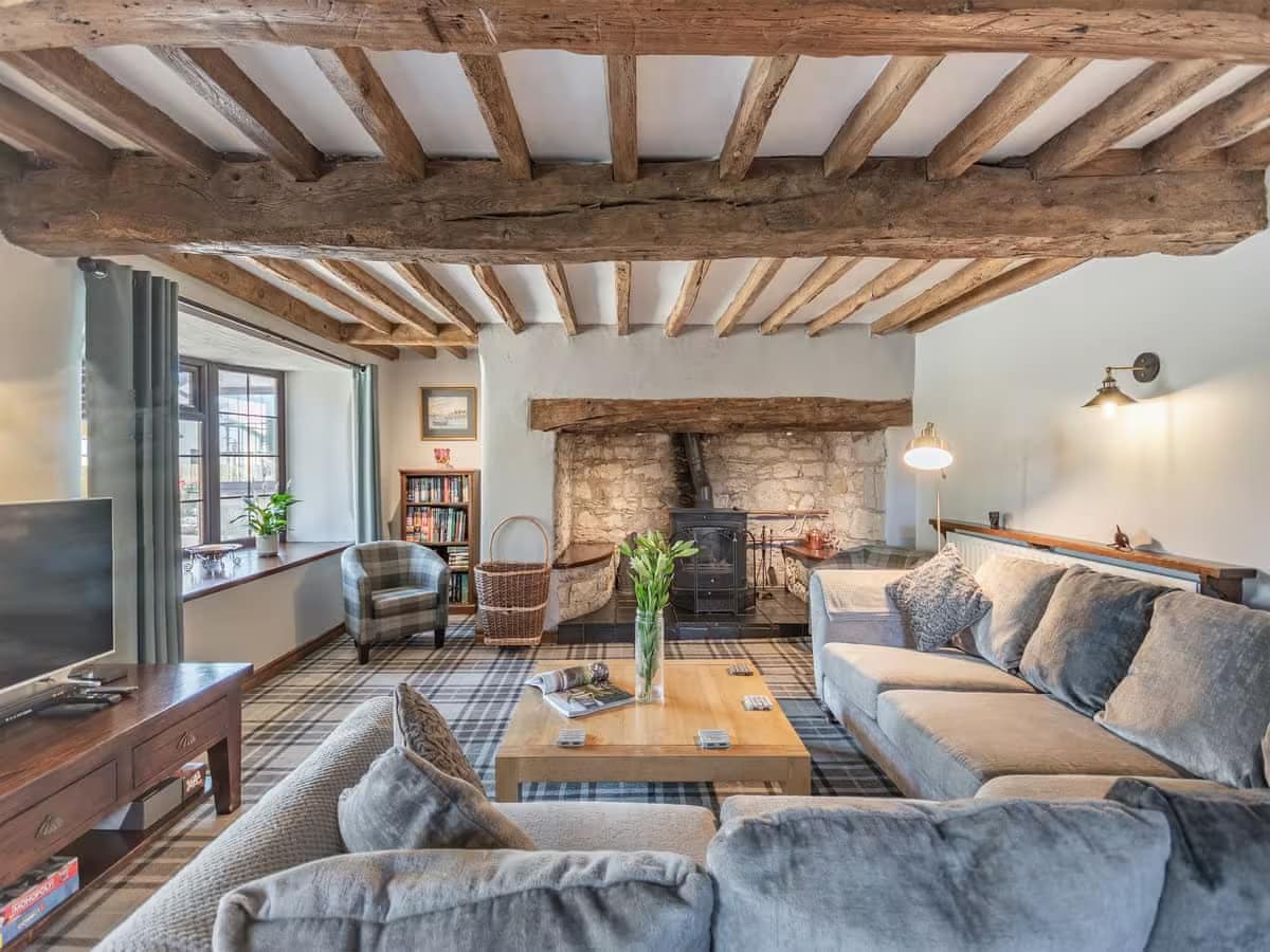 A cozy living area showcases exposed wooden beams and stone walls, featuring a large, comfortable sectional sofa. A wooden coffee table sits at the center, while a wood burner offers warmth. Natural light enters through a window, complemented by a bookshelf and decorative elements.
