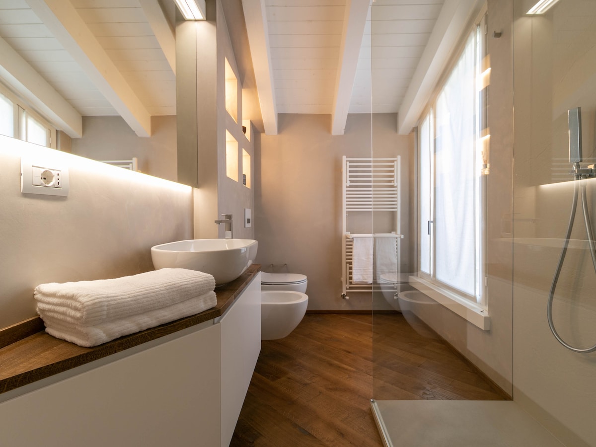 A modern bathroom is showcased with wooden accents and a spacious layout. A large shower with glass partition is present, alongside a sleek sink and bidet. Towels are neatly stacked on a wooden surface, while natural light filters in through large window panes.