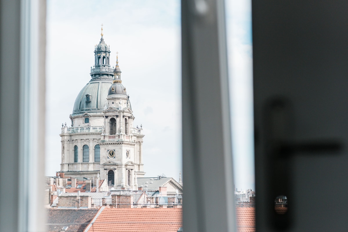 A view from the window showcases the intricate dome of St. Stephen's Basilica, framed by the window's sleek edges. The dome is adorned with detailed architectural features, set against a backdrop of a partially cloudy sky and the rustic rooftops of nearby buildings.