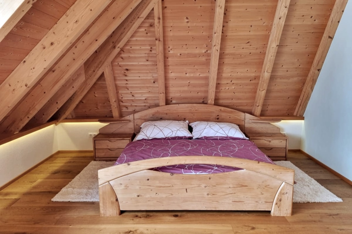 The sleeping area features a spacious wooden bed with a purple coverlet and white pillows. Wooden accents highlight the sloped ceiling, while soft lighting creates a gentle glow along the edges. A cozy rug is placed underfoot, enhancing the warm atmosphere of the room.
