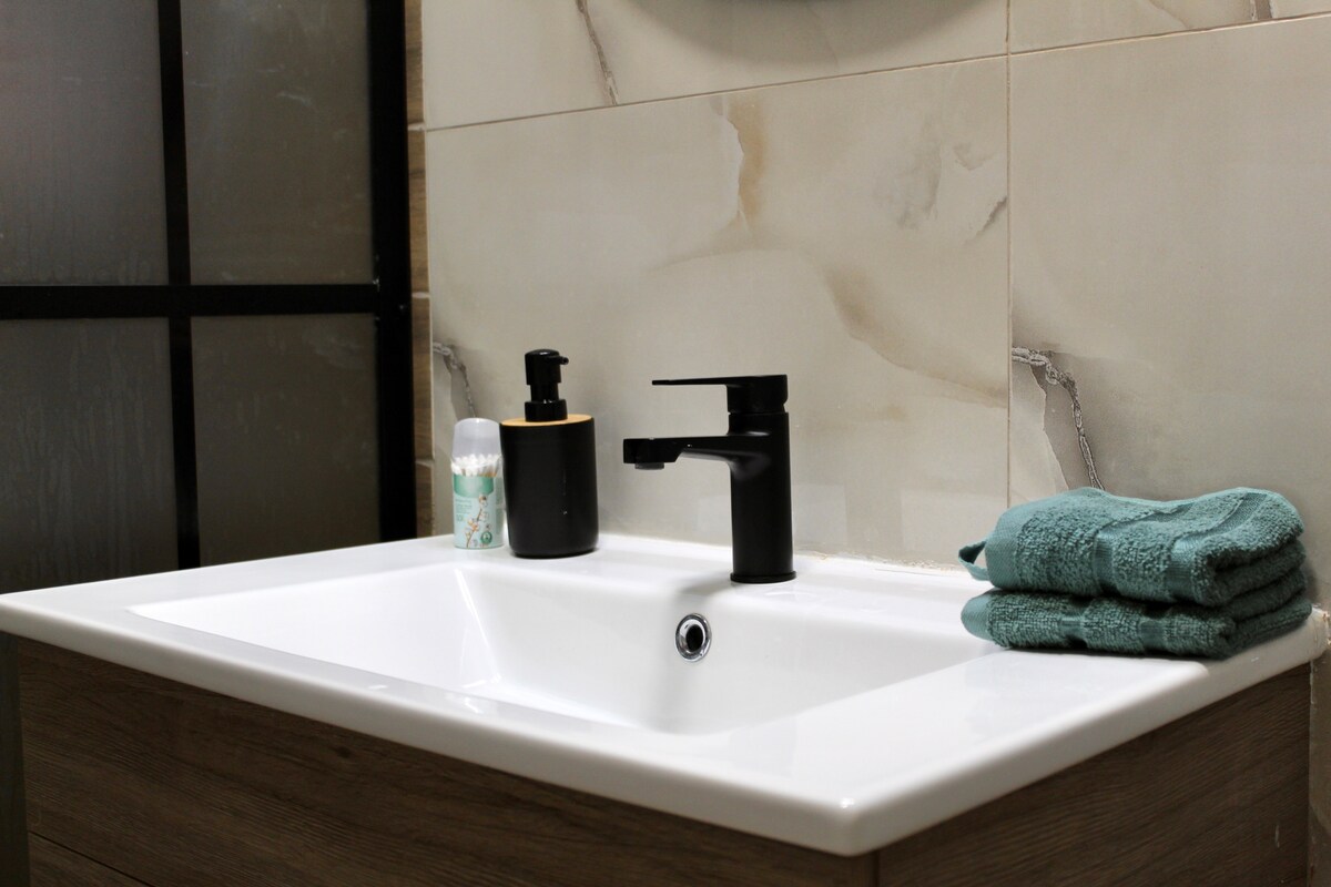 A clean bathroom sink area features a modern faucet with sleek lines. Complementing the sink are a black soap dispenser and a soft turquoise towel neatly folded beside it. The calming tiles provide a neutral backdrop.