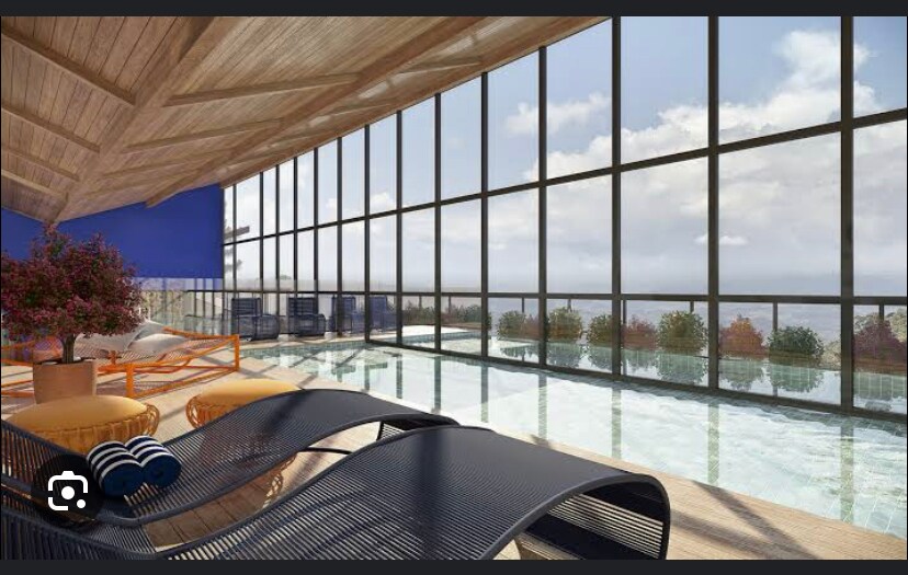An indoor pool area features large floor-to-ceiling windows, allowing natural light to illuminate the space. Relaxing lounge chairs are positioned along the pool's edge, providing a view of the surrounding greenery. Soft blue walls complement the wooden ceiling structure.