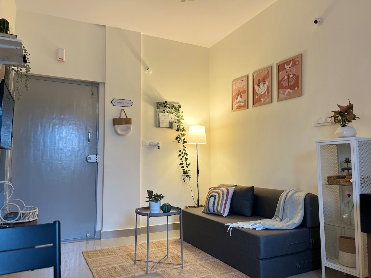 An inviting living area features a modern gray sofa, accentuated by decorative pillows. A small round table rests on a woven area rug, complemented by a floor lamp providing soft illumination. Two framed artworks and a plant add visual interest to the soothing, neutral-toned walls.