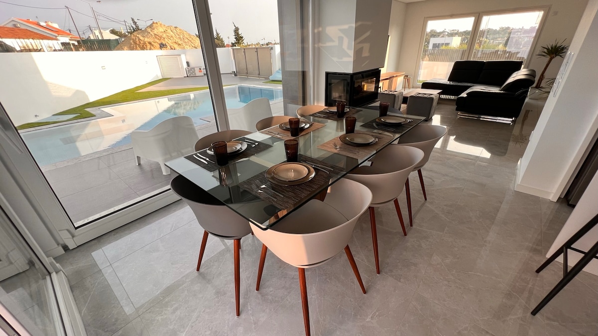 A dining area is featured with a glass table set for eight, accompanied by contemporary chairs with wooden legs. Large windows provide a view of the outdoor space, including a pool. Natural light fills the room, enhancing the clean, modern aesthetic.
