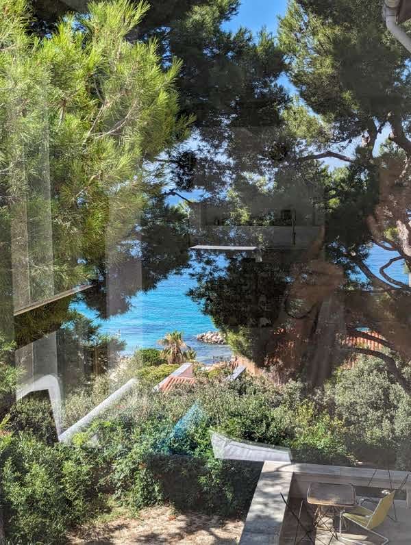 A view through glass showcases a vibrant blue sea framed by lush green trees. Clear skies are visible, complementing the natural landscape that descends towards the water. A section of a stone terrace is also seen, hinting at an outdoor seating area.