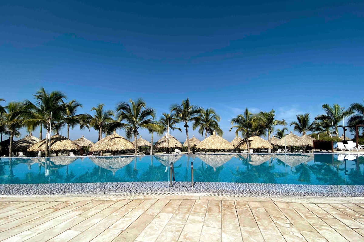A large pool reflects the clear blue sky, framed by palm trees and thatched umbrellas. The area features a beautifully tiled deck, providing ample space for relaxation around the pool. Lounge chairs are arranged along the edges, inviting guests to unwind.