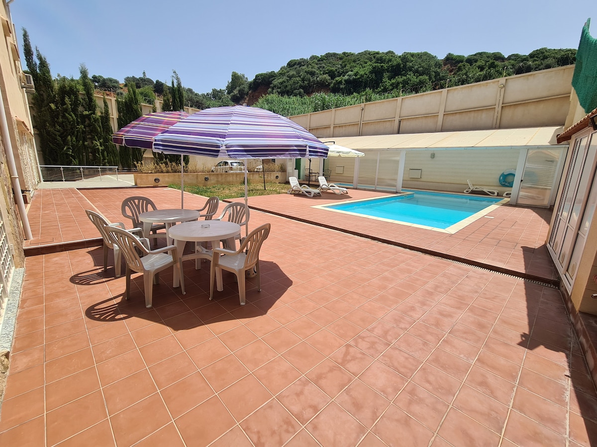 An outdoor area is equipped with a swimming pool and sun loungers. A shaded dining table with several chairs is situated nearby, offering a comfortable space for gatherings. The patio features tiled flooring, surrounded by greenery, providing a serene and spacious environment.