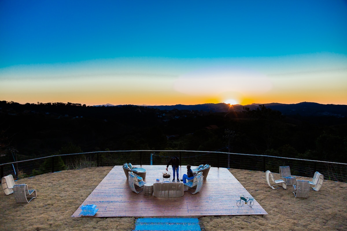 A spacious outdoor deck features comfortable seating arranged to enjoy panoramic views of the sunset. The sky transitions from blue to warm hues as the sun dips below the horizon, while surrounding hills create a serene backdrop.