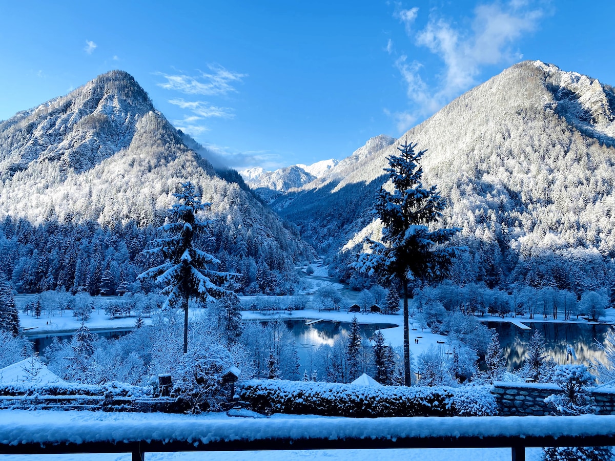 A snow-covered landscape reveals towering mountains and a serene lake, surrounded by frosted trees. The scene is illuminated by clear blue skies, providing a picturesque backdrop of winter beauty.