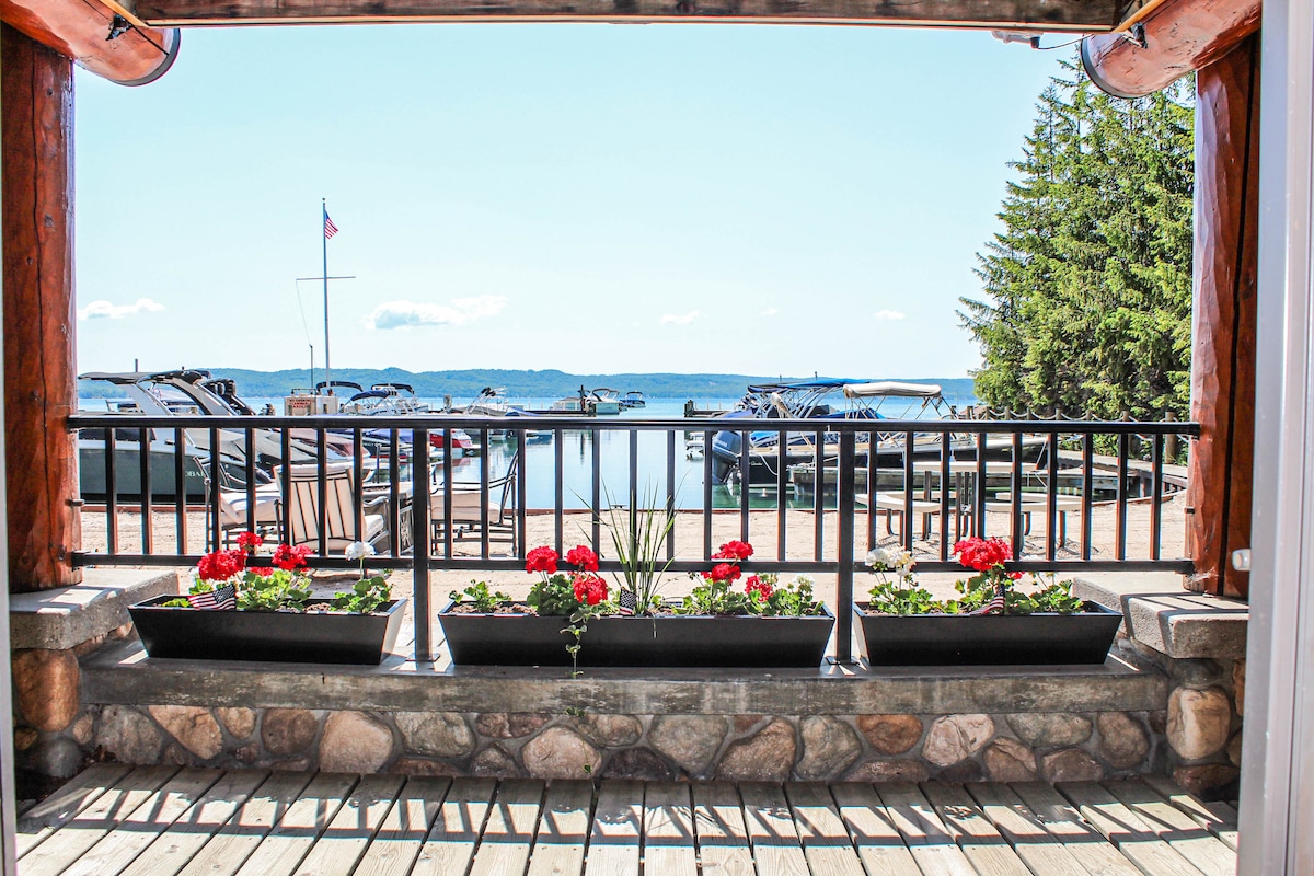 A view from a wooden deck features flower boxes with red geraniums, framed by rustic logs. In the backdrop, Glen Lake and boats are visible, with a flag fluttering gently nearby. Lush trees line the lakeshore, contributing to the serene lakeside ambiance.