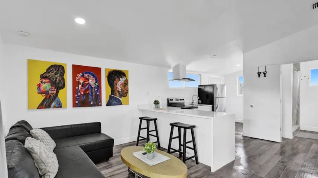 A modern living area features a dark sofa complemented by a light coffee table. Three colorful artworks adorn the wall, showcasing intricate portraits. Two bar stools are positioned at a kitchen counter with stainless steel appliances in the background, and natural light streams in from large windows.
