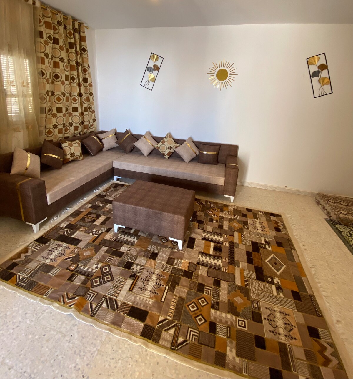 A comfortable living area is presented, featuring a sectional sofa adorned with accent pillows. A low coffee table is positioned atop a patterned area rug, while geometric wall art and a sun motif add visual interest to the neutral-toned space.