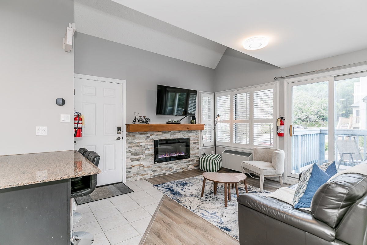 An inviting living area features a comfortable leather sofa and a cozy area rug, with a modern electric fireplace serving as the focal point. Large windows allow natural light to fill the space, and a sliding door opens to a private balcony.