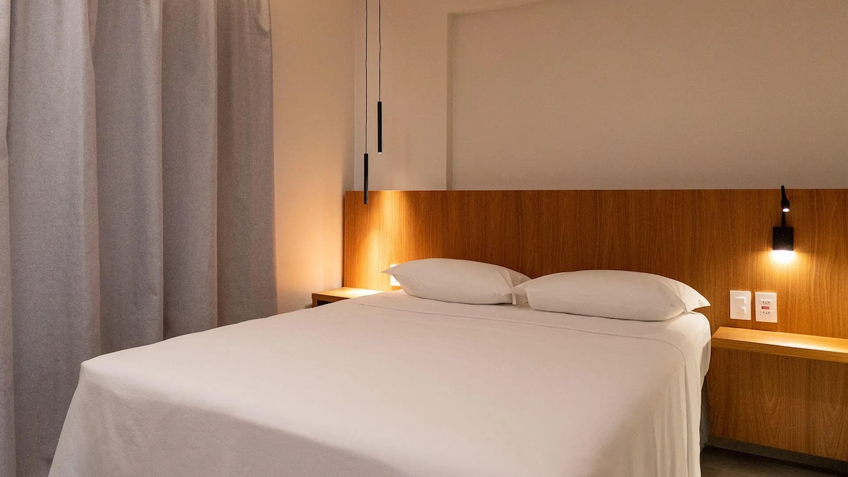 A spacious bed is positioned against a light wood headboard, softly illuminated by wall-mounted lamps. The surrounding walls are neutral, complemented by light gray curtains that add a calming touch to the atmosphere.