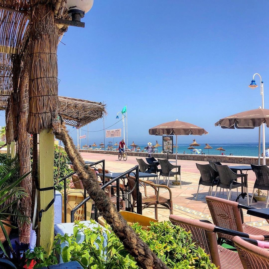 A vibrant outdoor dining area is showcased, featuring a variety of dining tables and shaded umbrellas. The view extends to the beach, with the ocean visible in the background. Green plants are placed throughout, adding a touch of nature to the setting.