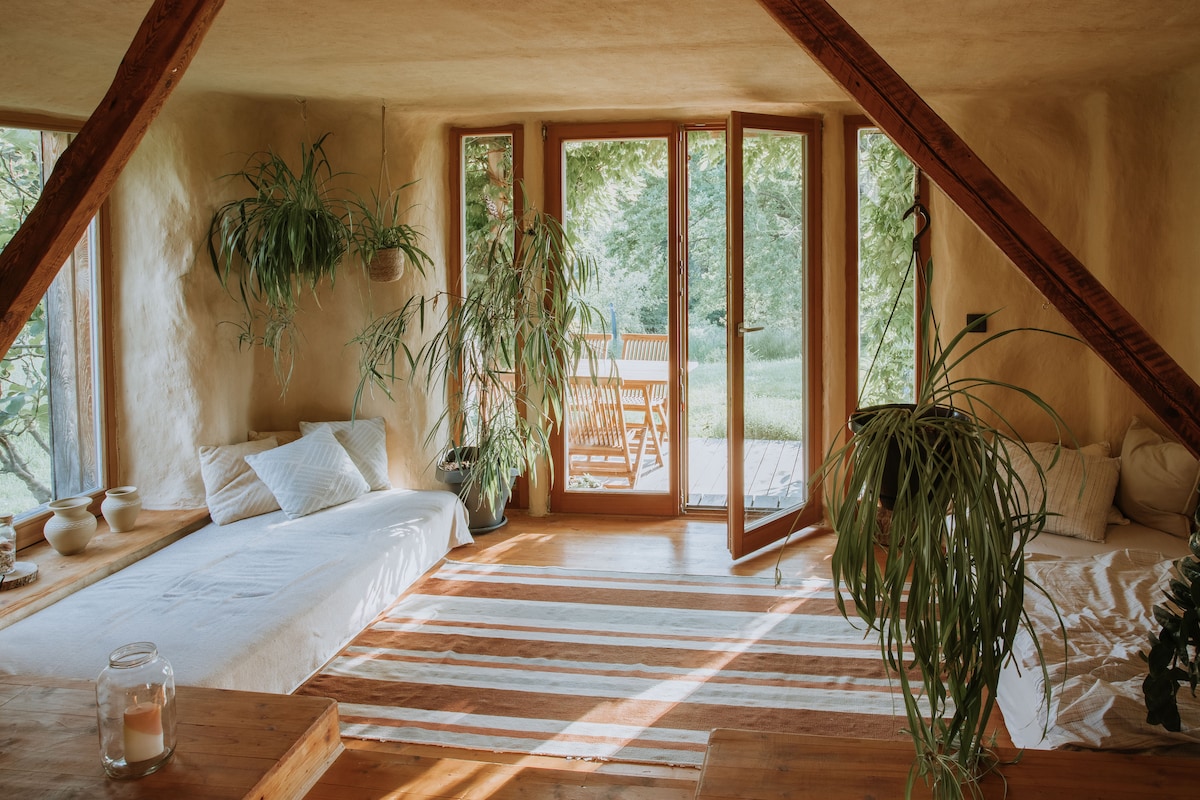 Natural light fills the spacious interior through large glass doors that open to the outside. The mud walls are complemented by natural wood beams. Potted plants adorn the corners, and a cozy sleeping area is arranged with soft linens on the wooden floor.
