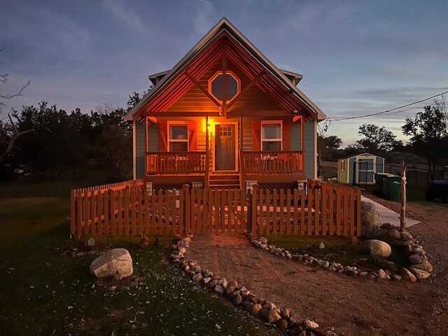 The exterior of a cottage is illuminated at dusk, highlighting the welcoming porch with a vibrant red gabled roof. A wooden fence surrounds the yard, and a path of smooth stones circles the manicured grass area, leading to the entrance.